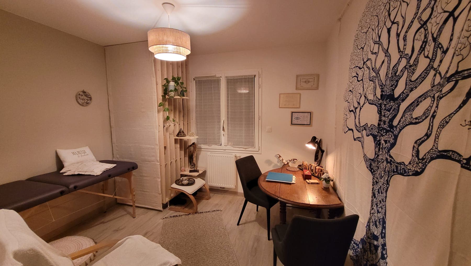 Le cabinet à Brax : table de soin avec un plaid, étagère en bois et plantes, bureau rond et tenture arbre de vie, sous une lumière tamisée.